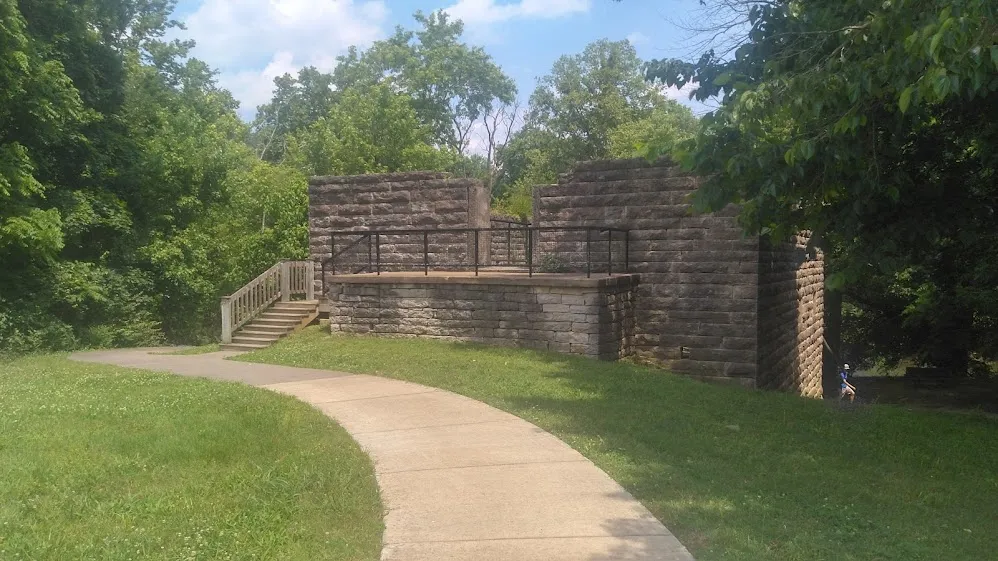 The stone walls of an old mill ruin beside a curving paved path and grass, framed by trees