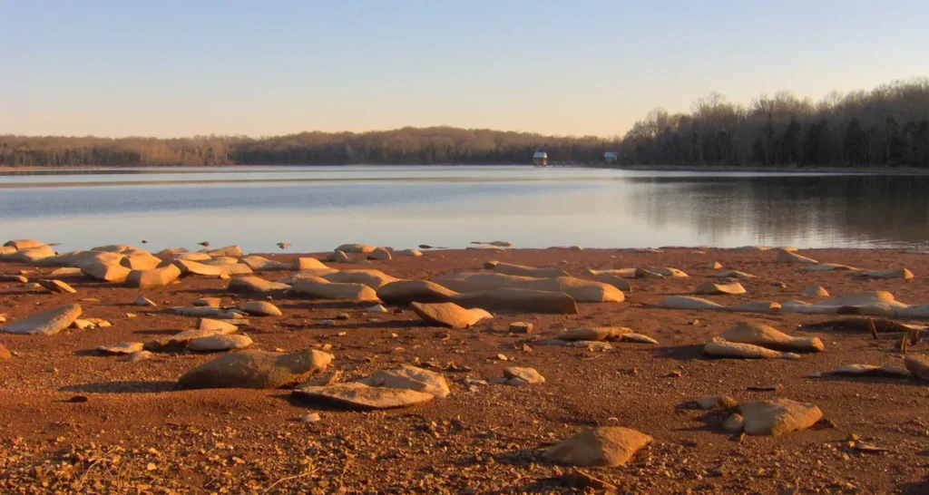 Flat rocks on a red-brown shore in low evening light, with a calm lake and a wooded far bank